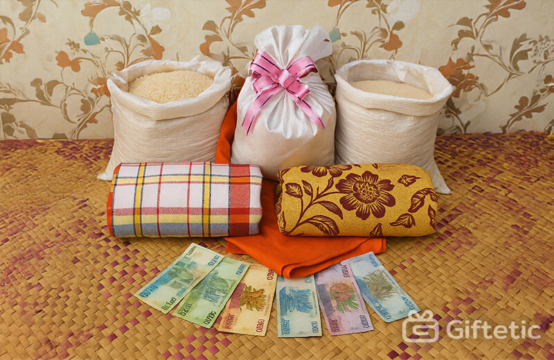 A display of traditional Malagasy Famadihana gifts arranged on a woven mat, including two open rice sacks, one white fabric gift sack tied with a pink ribbon, two colorful folded fabrics, and a spread of Malagasy banknotes. The setting features a patterned wall and symbolizes the offerings typically made during Madagascar’s Famadihana (turning of the bones) ancestral celebration.